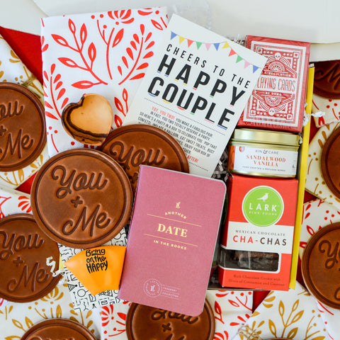Gift box on red background filled with leather coasters, date night passport, cookies, playing card, wood heart and happy couple card: Engagament Gift Idea