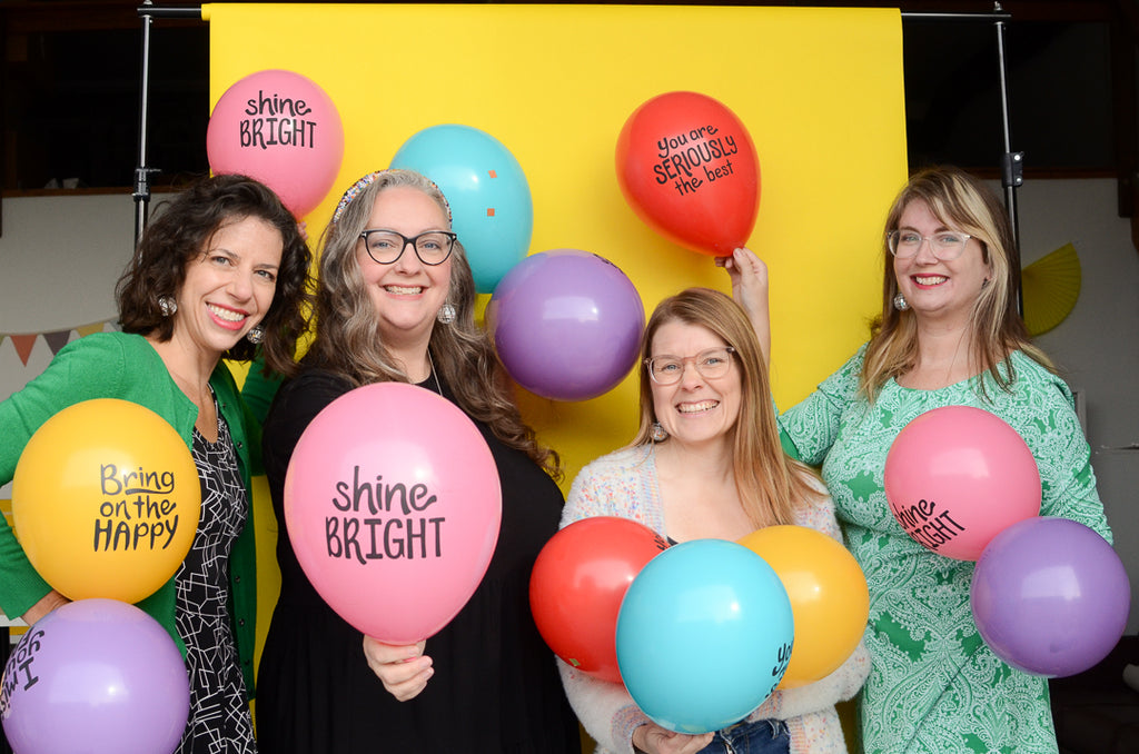 Photo of four women holding a rainbow of ballooons agaist a yellow backdrop