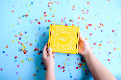 12 Best Ways to Celebrate Your Work-from-home employees with The Confetti Post Gift Box Company: Image of yellow gift box on a blue background with rainbow confetti scattered around