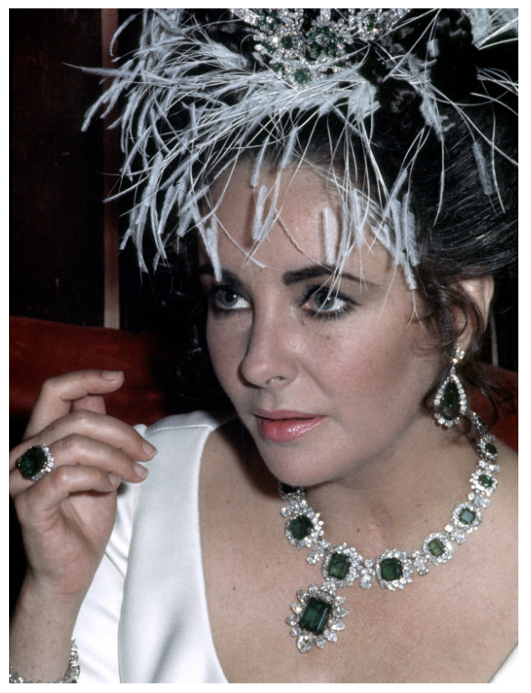 Color  photograph of Liz Taylor adorned with the intricate Bulgari emerald suite of a necklace, earrings and a ring, their elegance accentuated against her attire and glamorous demeanor