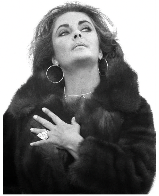 Black and white photograph of Elizabeth Taylor, clutching her fur coat close to her chest, prominently displaying the Taylor-Burton diamond on her finger with a look of pride and elegance