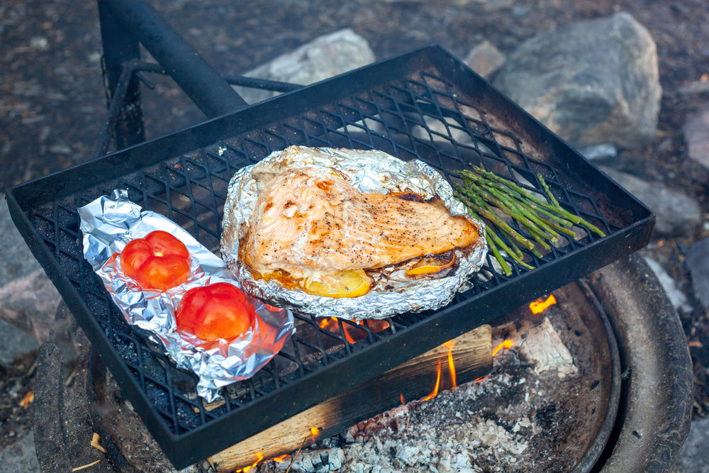 Sliced tomato on foil, fish with lemon on foil, and asparagus, all on a grill over an open fire