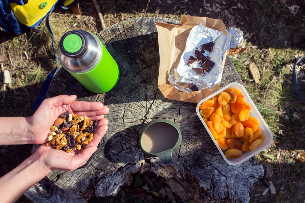 Various camping snacks: Dried apricot in a tub, nuts in cupped hands. and dark chocolate on foil