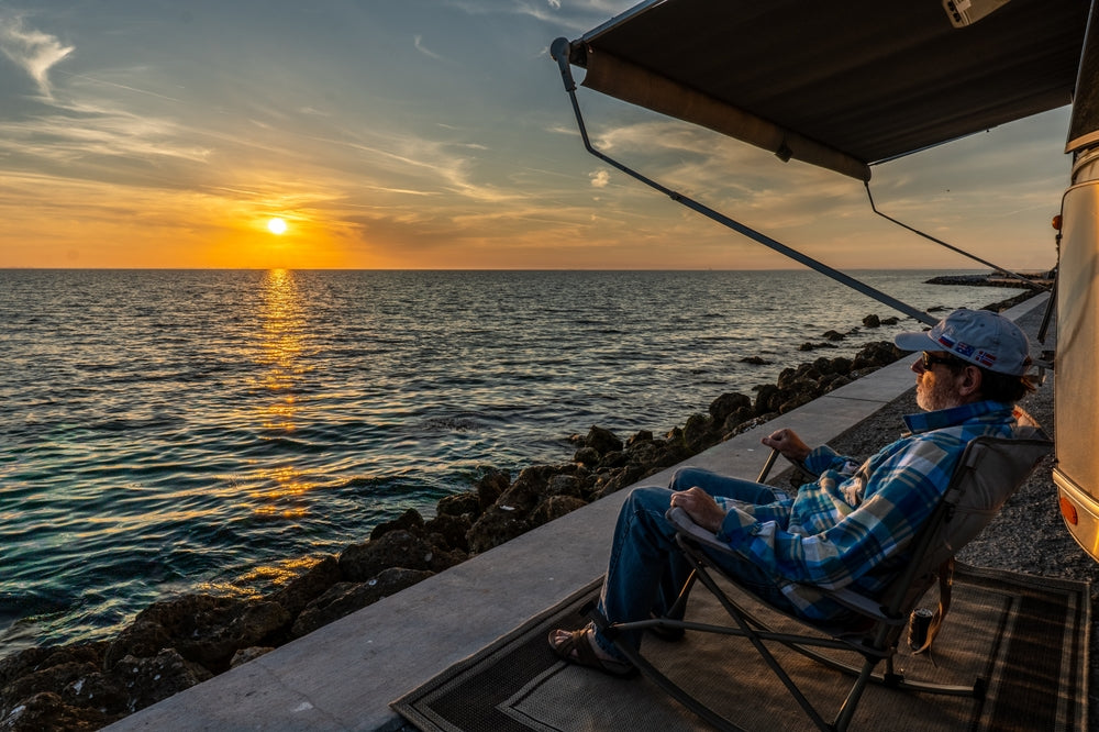 Man sitting under camper trailer awning by the water during sunset 