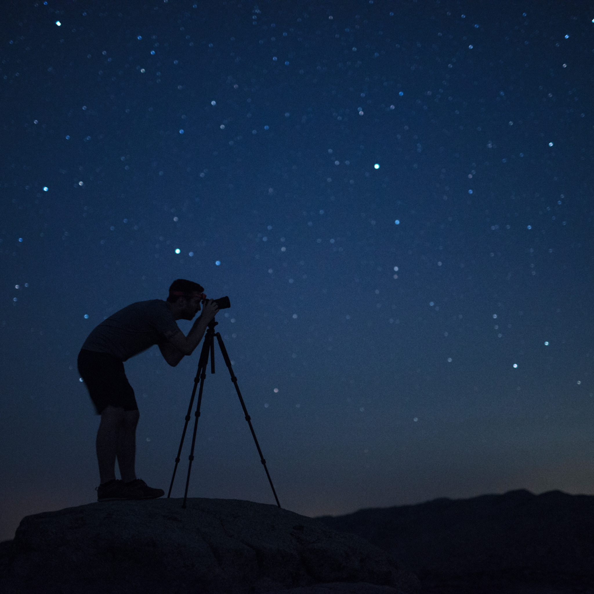 Man bending over looking into his camera set up on its tripod on a mountain taking a photo of the night sky