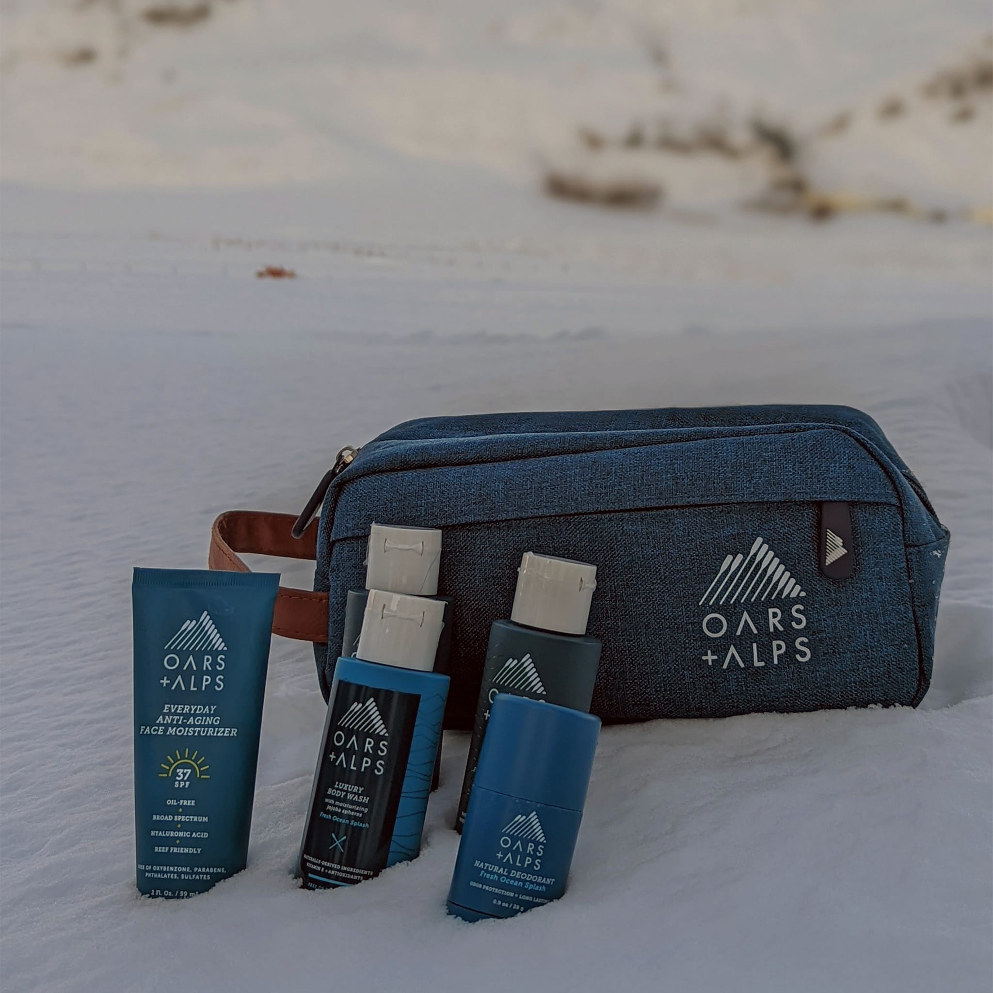 Oars + Alps dopp kit, Everyday Anti-Aging Face Moisturizer with SPF 37, Aluminum-Free Deodorant, and Travel Size Shampoo, Conditioner, and Body Wash in the snow in front of a mountain