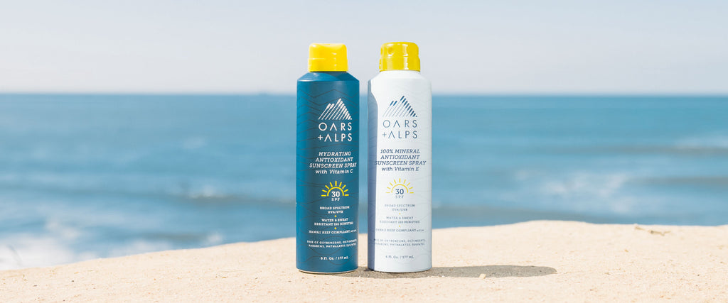 Oars + Alps SPF 30 Spray Sunscreen and Mineral Sunscreen bottle in the sand on a beach.