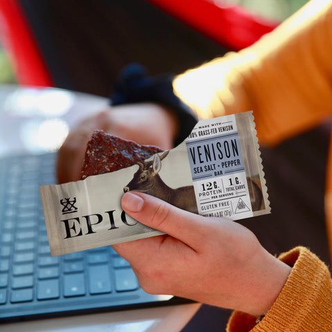 A close up of a woman holding an EPIC Venison Sea Salt Pepper Bar while working on a laptop.