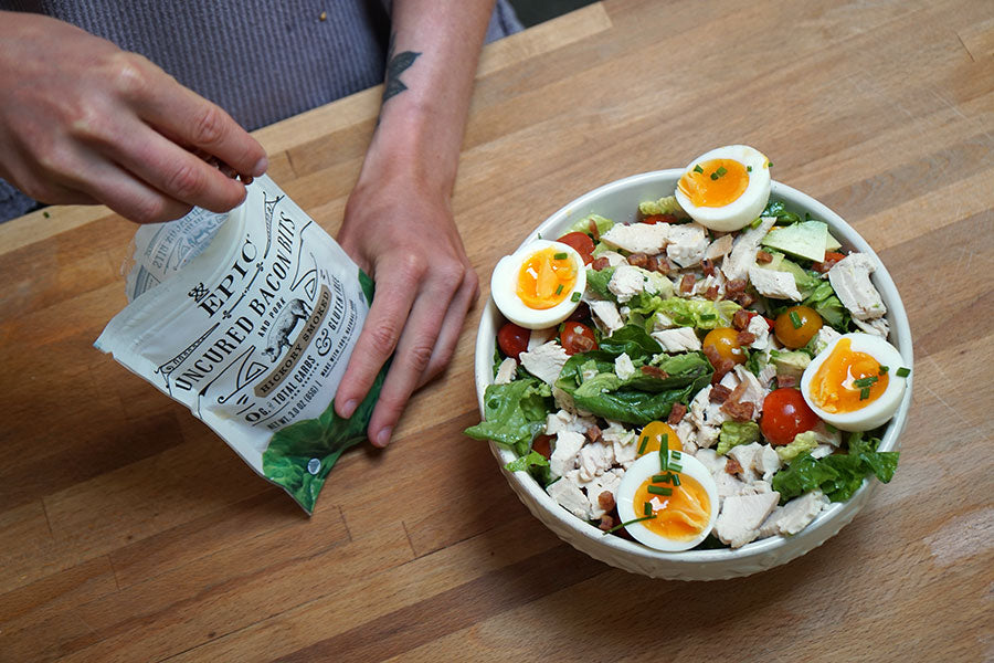 A complete Summer Cobb Salad with an individual adding EPIC Hickory Smoked Topper Bits on top of the salad.