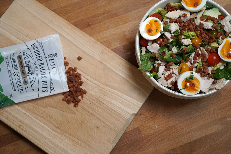 Image of a Summer Cobb Salad next to a cutting board with a bag of EPIC Hickory Smoked Bacon Bit's laying on top of it.