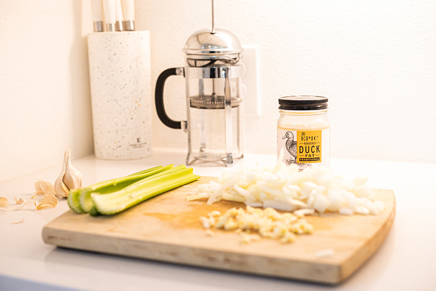 A cutting board with celery stalks and chopped onion on top of it. Next to the cutting board is garlic cloves, EPIC Duck Fat and a French press.