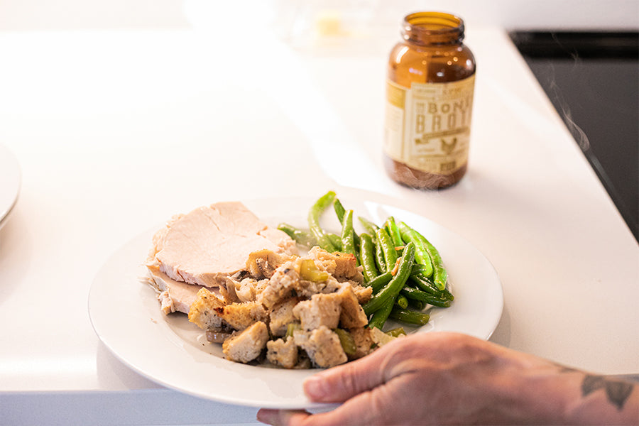 A plat of turkey, green beans, and EPIC's Bone Broth dressing being grabbed from the counter next to a bottle of EPIC Turkey Bone Broth.