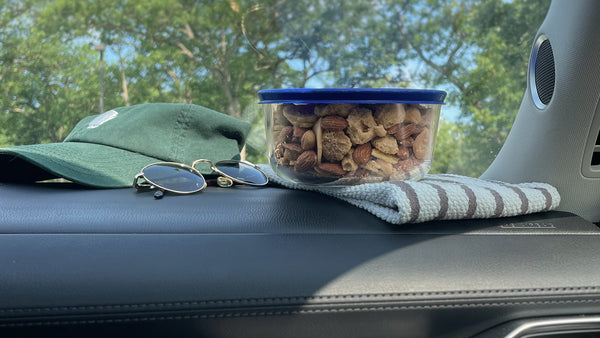 A dashboard of a car with a hat, sunglasses, and container of EPIC's Grain-Free Maple Bacon Snack Mix sitting on it.