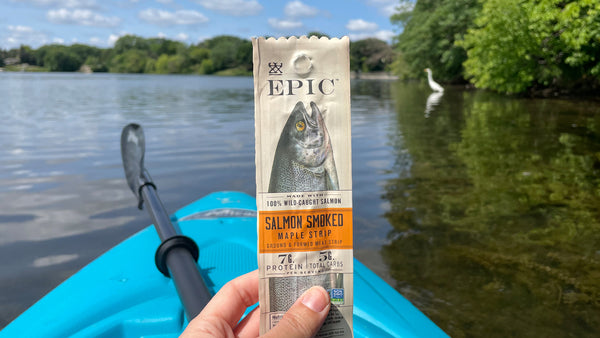 A close-up image of a person holding EPIC's Smoked Salmon Maple Snack strip on a kayak with a lake in the background.