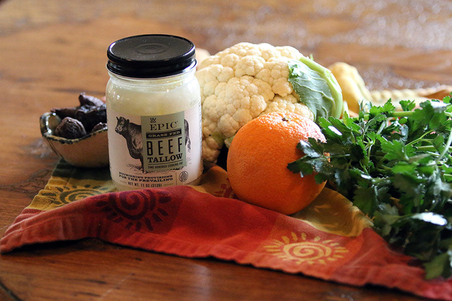 Head of cauliflower, jar of Epic Provision's Beef Tallow, a handful of arugula, a jar of apricots and an orange arranged on a kitchen towl.