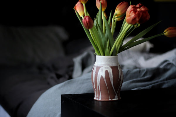 a wheel thrown pottery vase filled with tulips glazed with a splash of cream glaze sits on a table at the foot of an unmade bed.