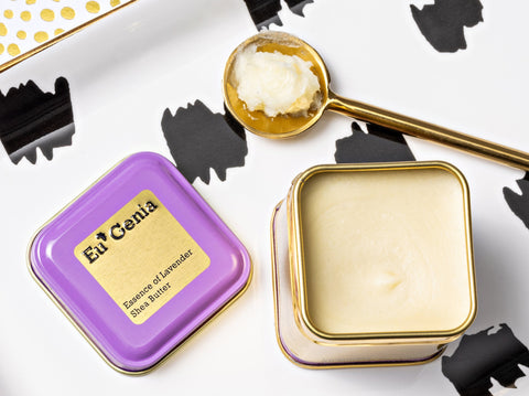 Open purple tin of shea butter next to a gold scoop with shea butter both on a white tray with black splotches 