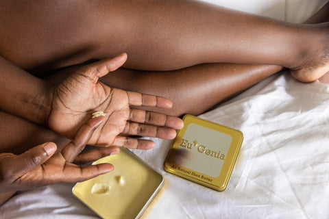 Person putting shea butter into the palm of their hand, open tin of shea butter in the background with bare legs visible.