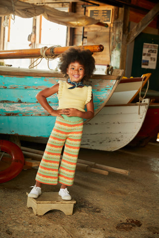 African American Model about the age of 5 is wearing the Fiora hand knitted vest by the child;s designer Misha and Puff.  She is leaning against a vintage green fishing boat while wearing stripped wide legged pants.  Shop Taylorandmax.com for unique and special clothes for your girl or boy.