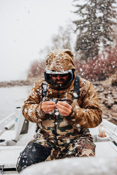 In this image, from Moonshine Rods, an angler is tying a fly on his line while it's snowing.