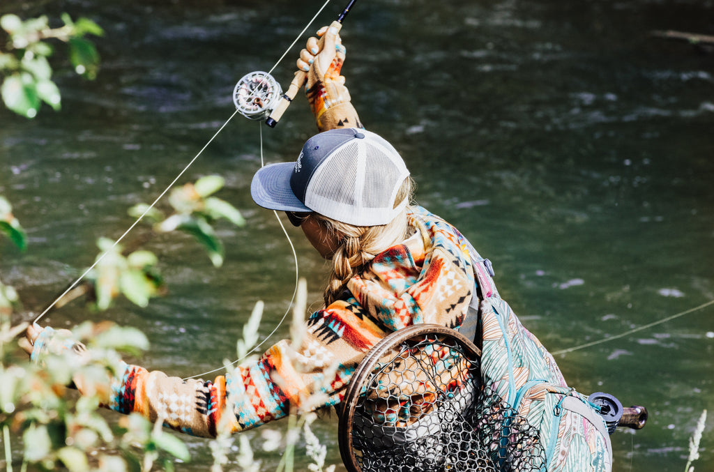 In this image, from Moonshine Rods, a female angler is casting a fly rod into a river.