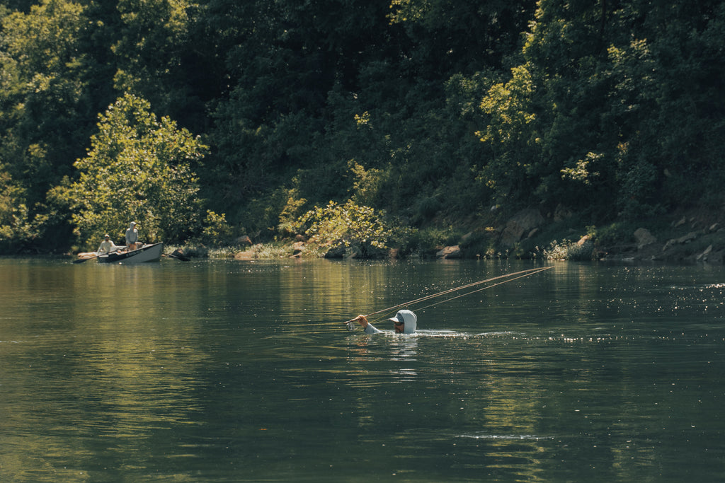 In this image, from Moonshine Rods, an angler is swimming across a river with his fly rod in the air.
