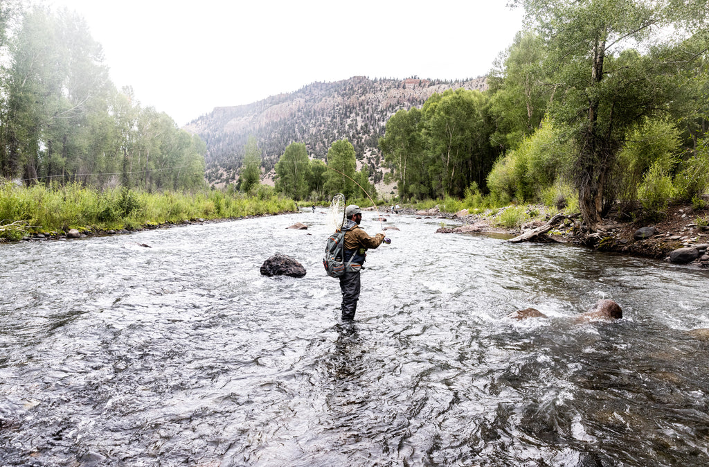 In this image, from Moonshine Rods, a fly angler is casting a rod while standing in the middle of a river.