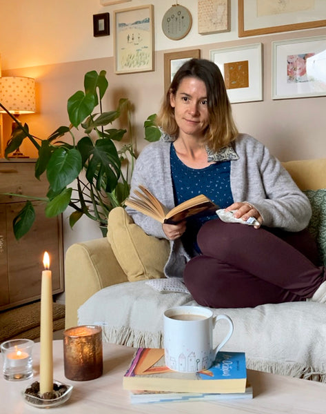 Charlotte Macey at home on the sofa in a cosy living room with candles, hand warmers, a wheat heat bag and a book.