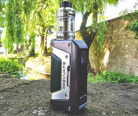 A Geekvape Aegis Legend 2 vape kit stood on a concrete step, with trees and brick walls in the background.