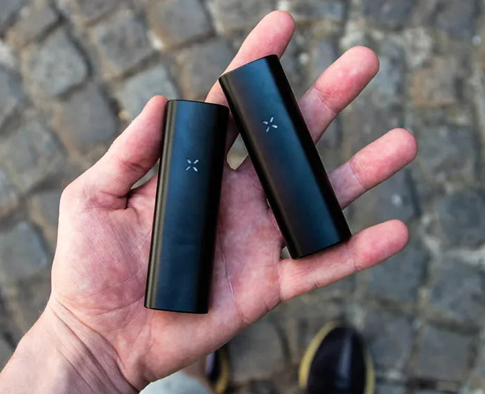 The PAX Mini and PAX Plus held in someones hand over a cobbled street.