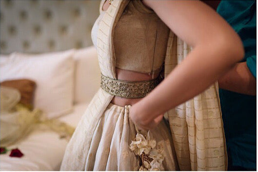 An Indian bride getting dressed for her wedding. She is wearing a handwoven silk lehenga set in gold. The photograph captures the embroidered details of her belt.  