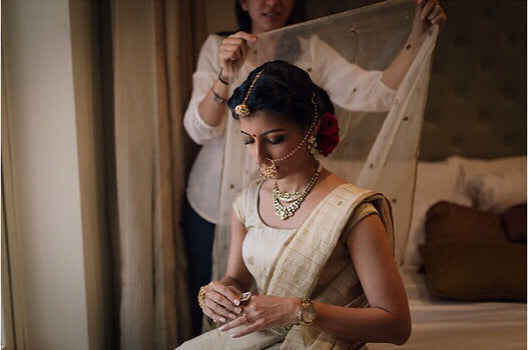 An Indian bride getting ready for her wedding ceremony. She is dressed in a handwoven silk benarasi lehenga outfit. A woman is helping her put her veil on the head.