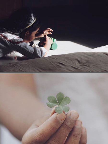 A collage of two images part of a photo story on memories of childhood by photographer Nirmala Mayur Patil. The top image is of a little girl lying on her tummy in a spot of sun on a bed playing with a green wooden car. The bottom image is of a child's hand holding a clover leaf.