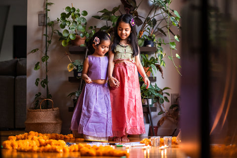 Indian sisters celebrating the festival of Diwali together creating a decoration with marigold flowers and colour dressed in traditional Indian attire of lehenga and choli in beautiful handwoven cotton silk textiles from Love the World Today