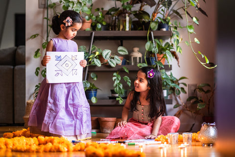 Younger sister showing her rangoli drawing to her older sister while they are busy prepping for Diwali - an Indian festival of lights. The sisters are dressed in beautiful colourful Indian festive wear and surrounded by marigold flowers and earthen lamps