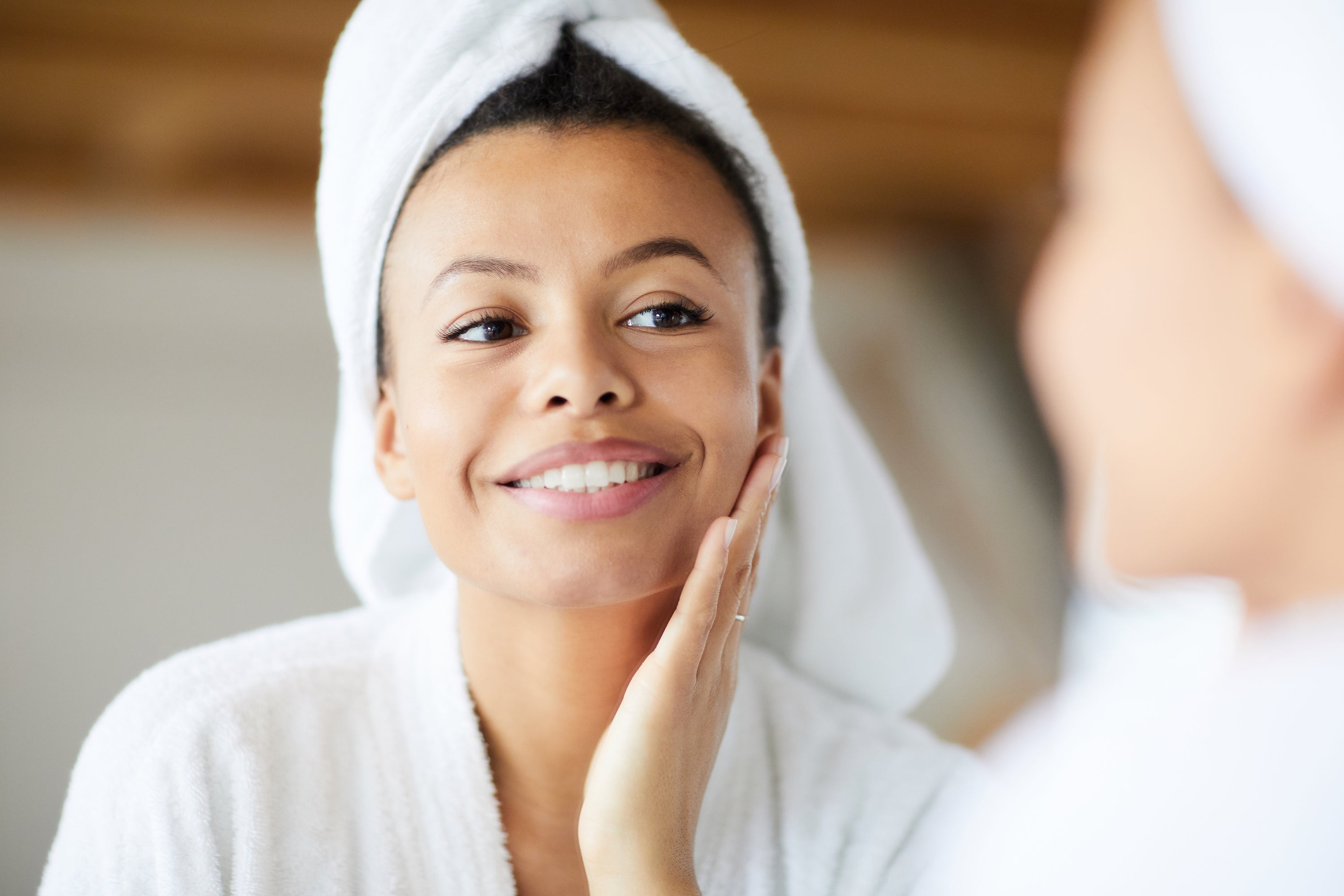 woman of color with clear skin touching cheek and looking in mirror with towel on head wearing bathrobe