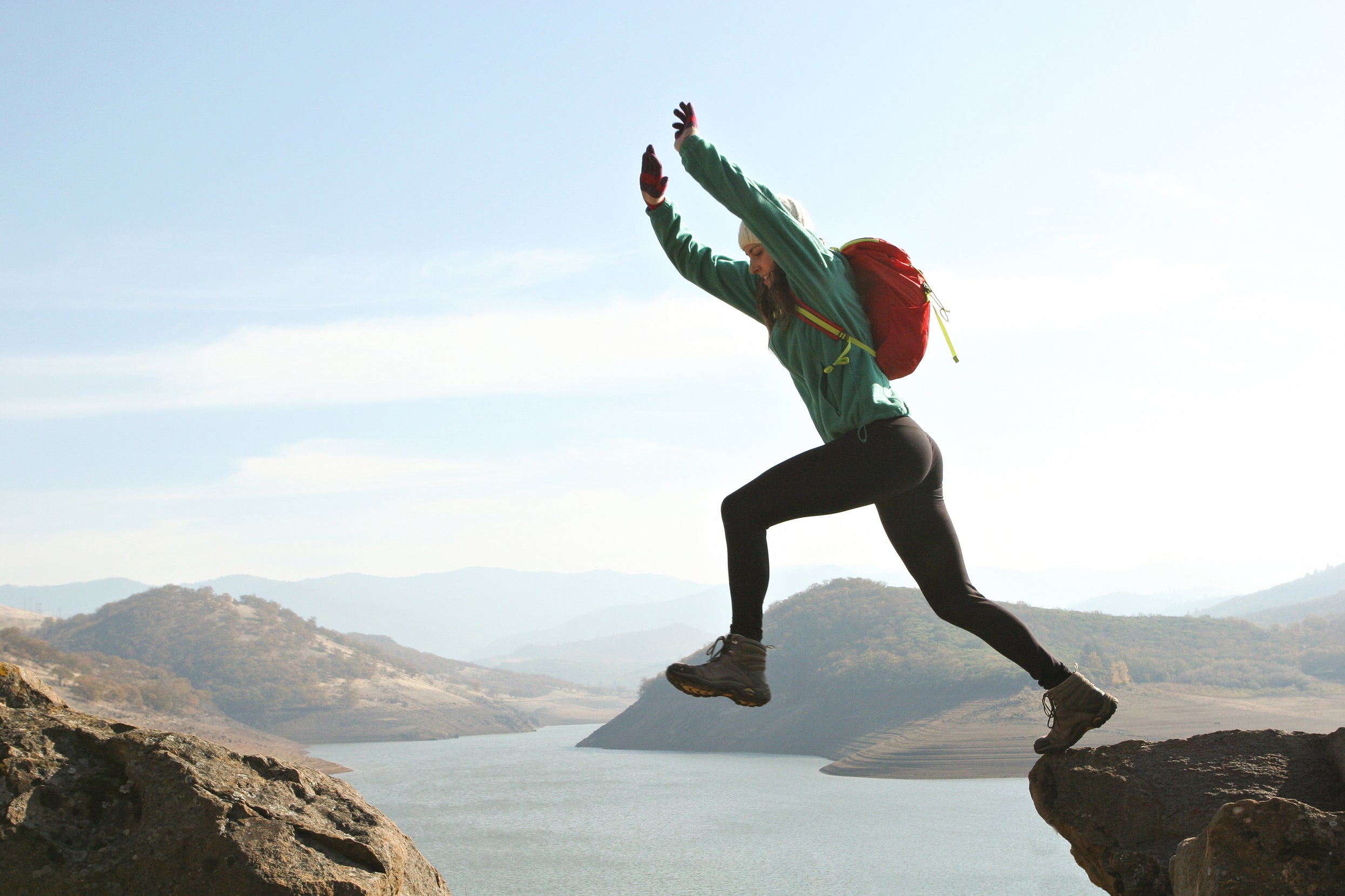 female hiker with backpack taking leap between two rocks with lake in background