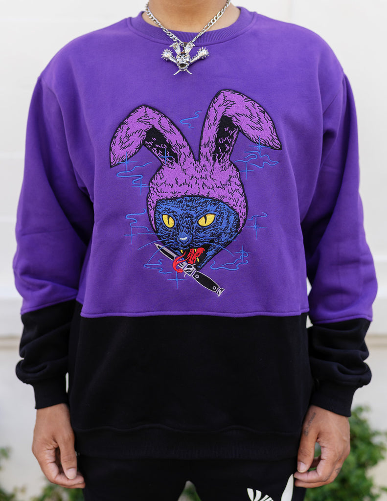model wearing half purple and black crew neck sweatshirt with an embroidered cat with bunny ears and a knife in its mouth design