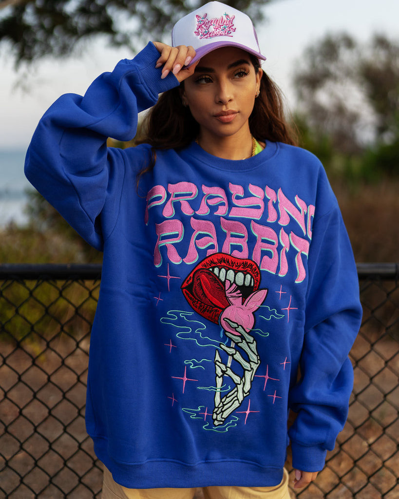 model wearing blue crew neck with a large embroidered skeleton hand holding a rabbit lollipop with lips licking it design