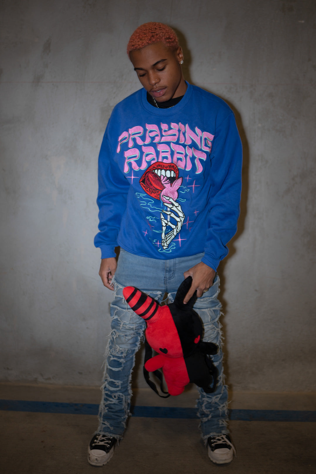 male model wearing candy blue embroidered crew neck with a design that shows a skeleton hand holding a rabbit lollipop. the model is also holding a red and black bunny plush