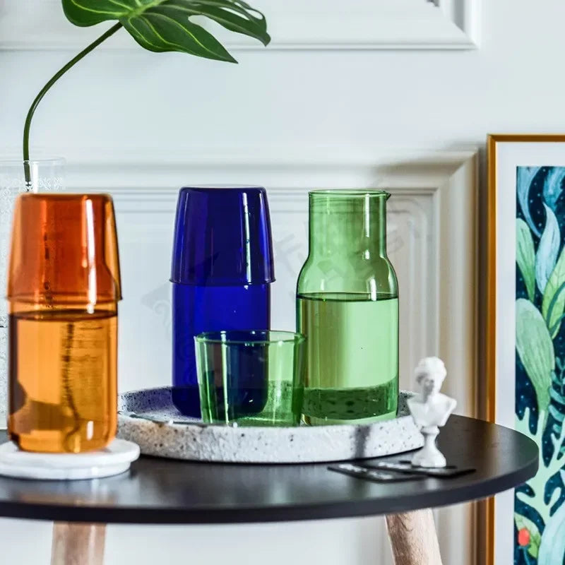 Three colorful glass vases on a table with a plant and framed picture in the background.