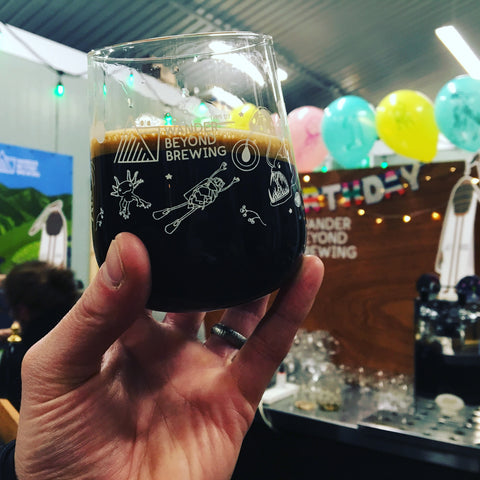 Hand holding a glass of dark beer with multicoloured balloons in the background.
