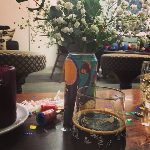 A glass of dark beer alongside flowers in a beer can and party streamers.