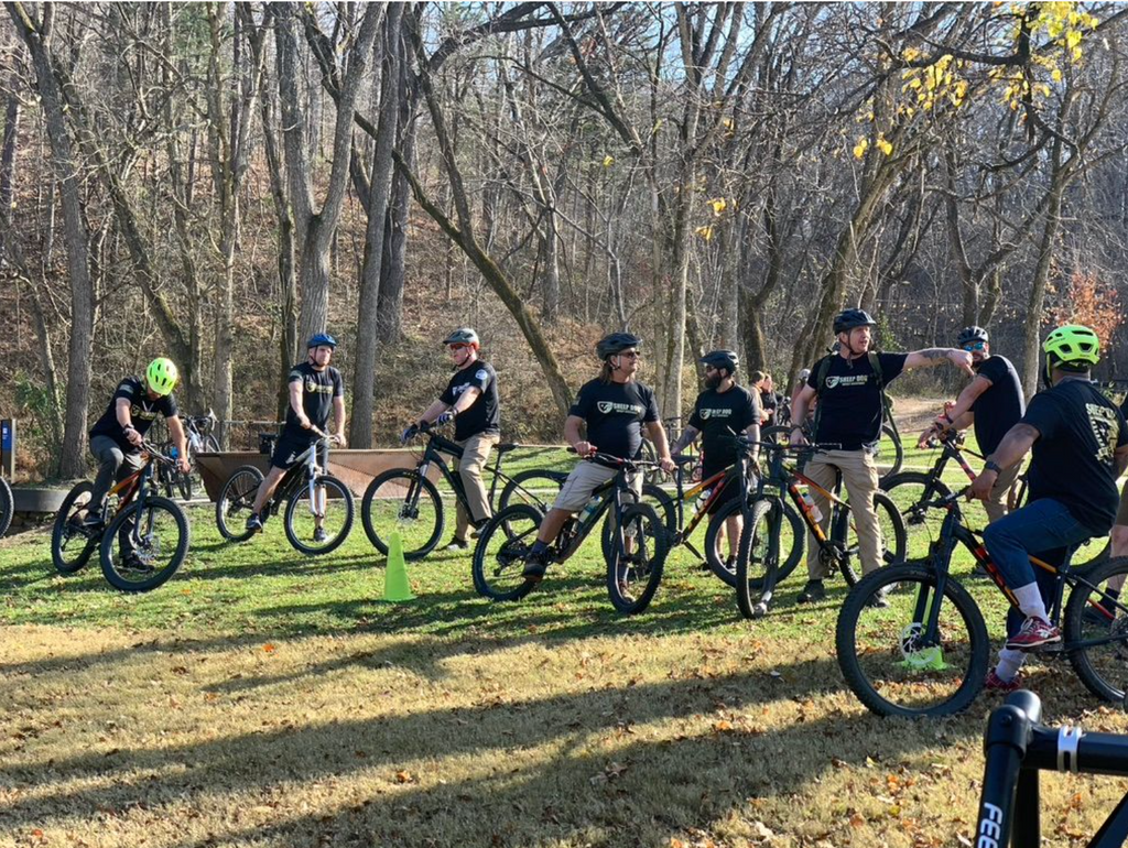 Sheep Dog Bike Ride - many veterans gather wearing bike helmets with their bikes. There is green grass and trees in the background.