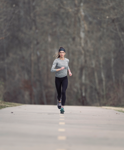 Woman runs toward the camera, there is woods behind and she is on a path