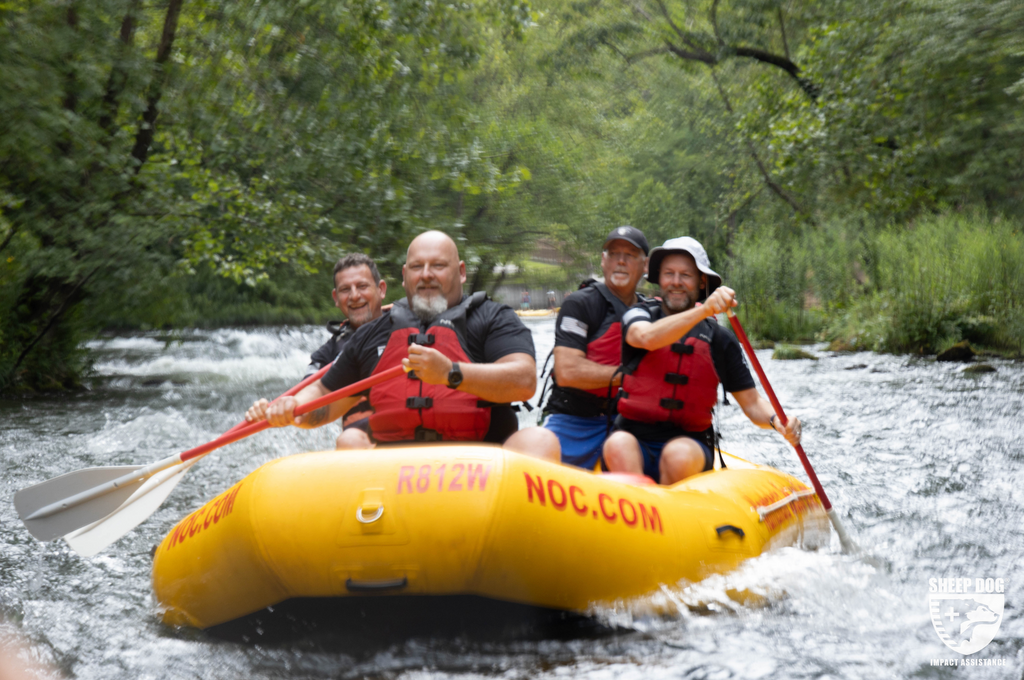 Veterans on a river outdoor adventure with Sheep Dog, the white man in the front is bald with a beard, and the man behind him in his raft has short hair, the men on the other side are wearing a hats