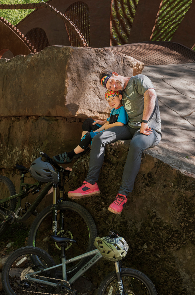 A father and son sit on a large rock formation wearing biking gear and their bikes and helmets are below them.