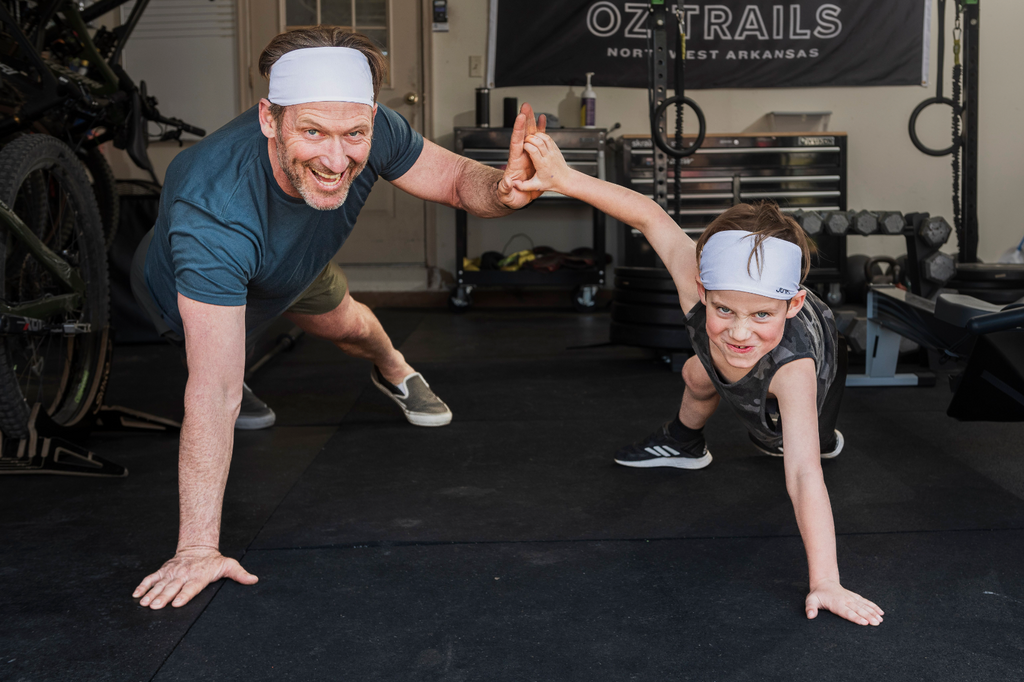 A father and son do pushup high fives in their garage gym. They are wearing JUNK headbands and smiling at the camera.