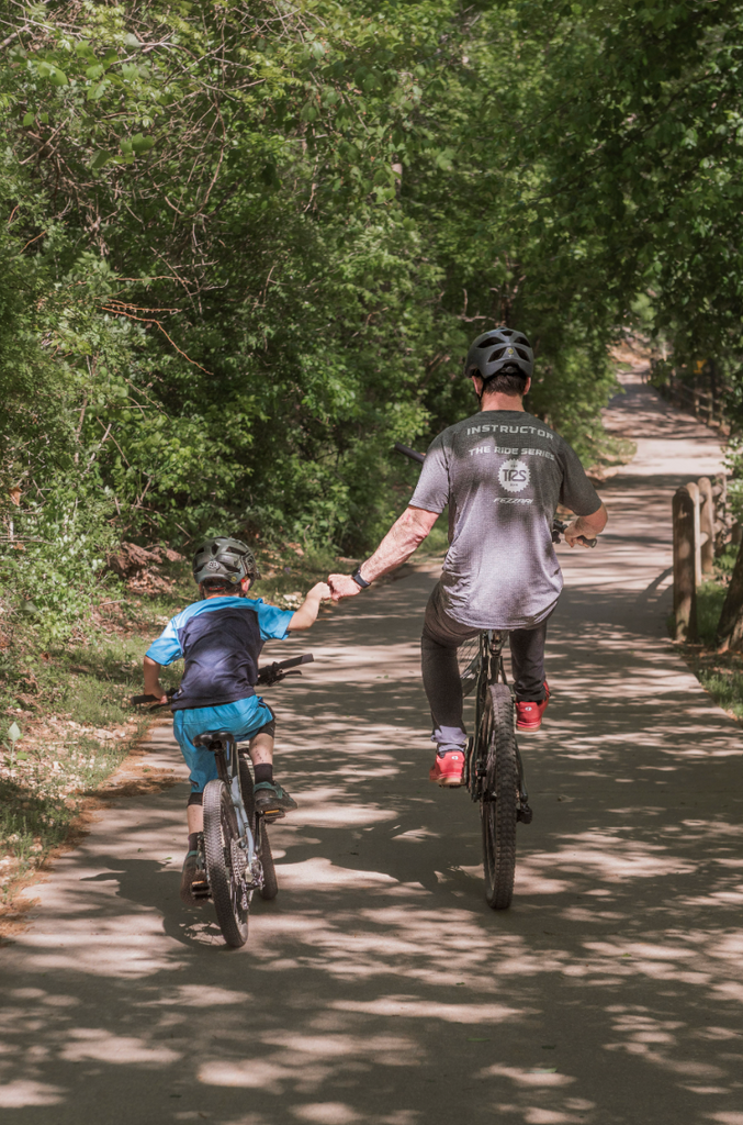 A father and son ride away from the camera on a trail in the woods on their mountain bikes and they give each other a fist bump.