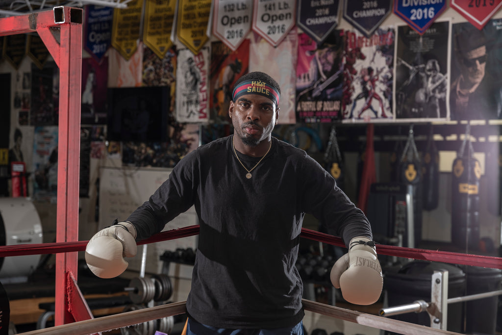 Kalvin stands in the corner of the ring with his arms resting on the ropes. You can see his gym behind him. There are banners and posters behind him. He is wearing a “hot sauce” junk headband. 